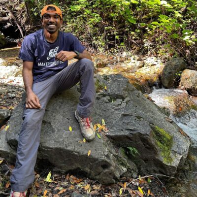 A color photo of a man sitting on a large rock, by a stream.