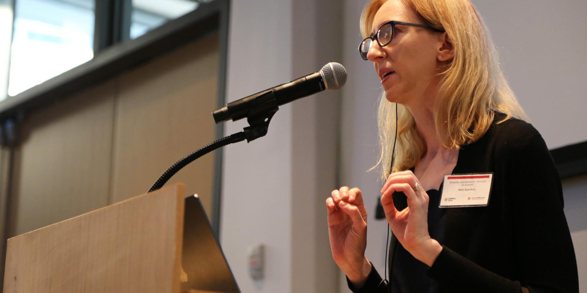 A color photo of a woman speaking at a microphone.