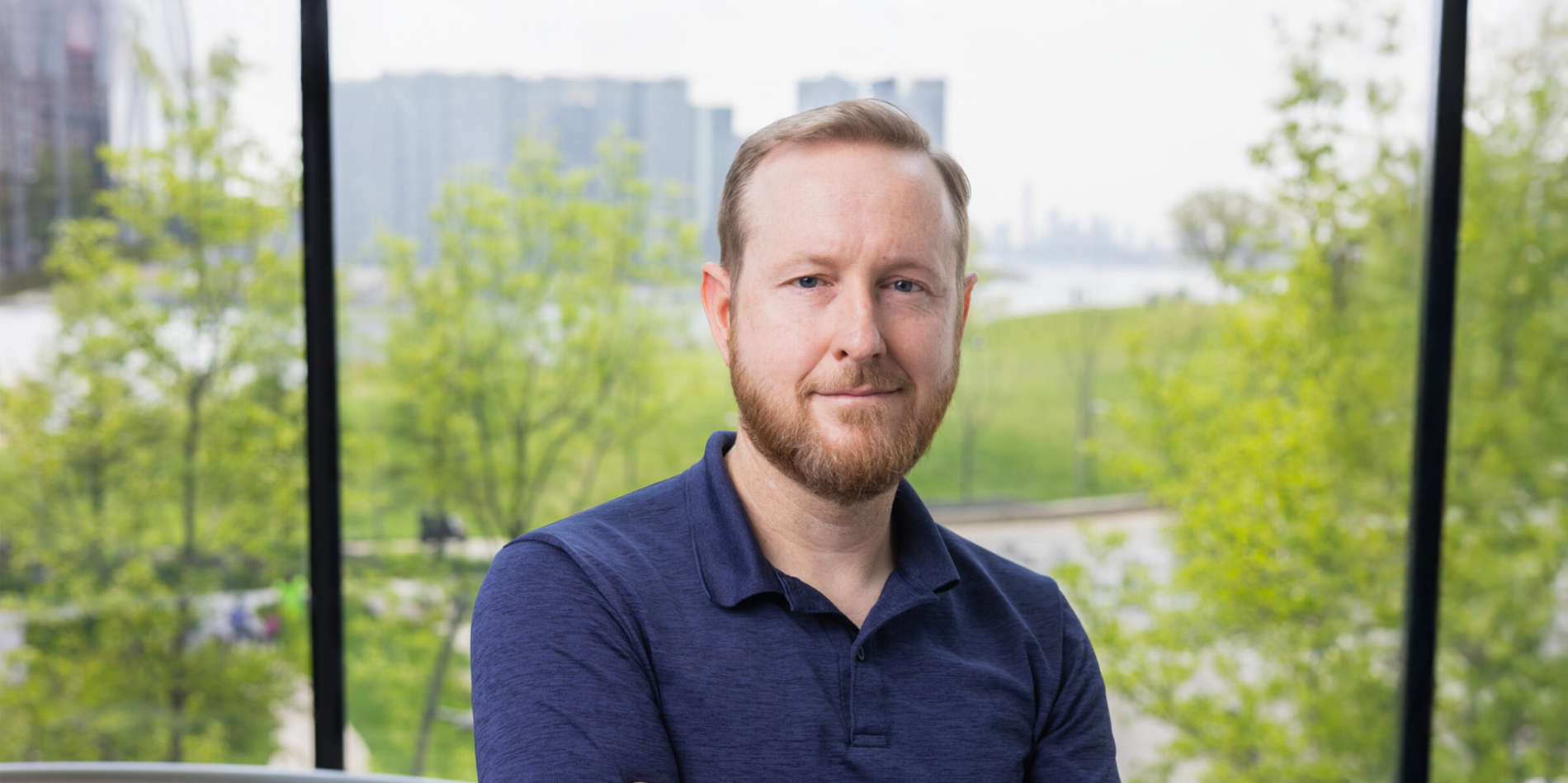 A color photo of a man with a beard standing front of a window for a photo.