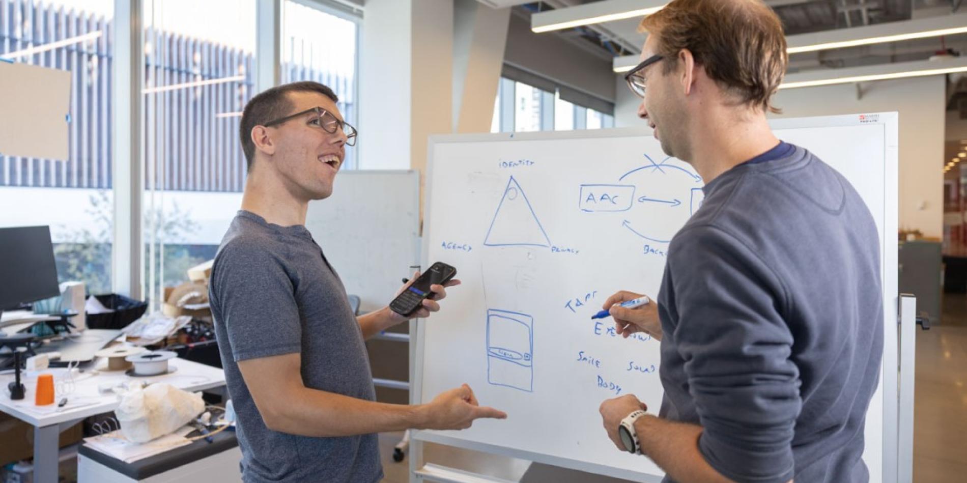 A color photo of 2 men discussing a project in front of a white board.