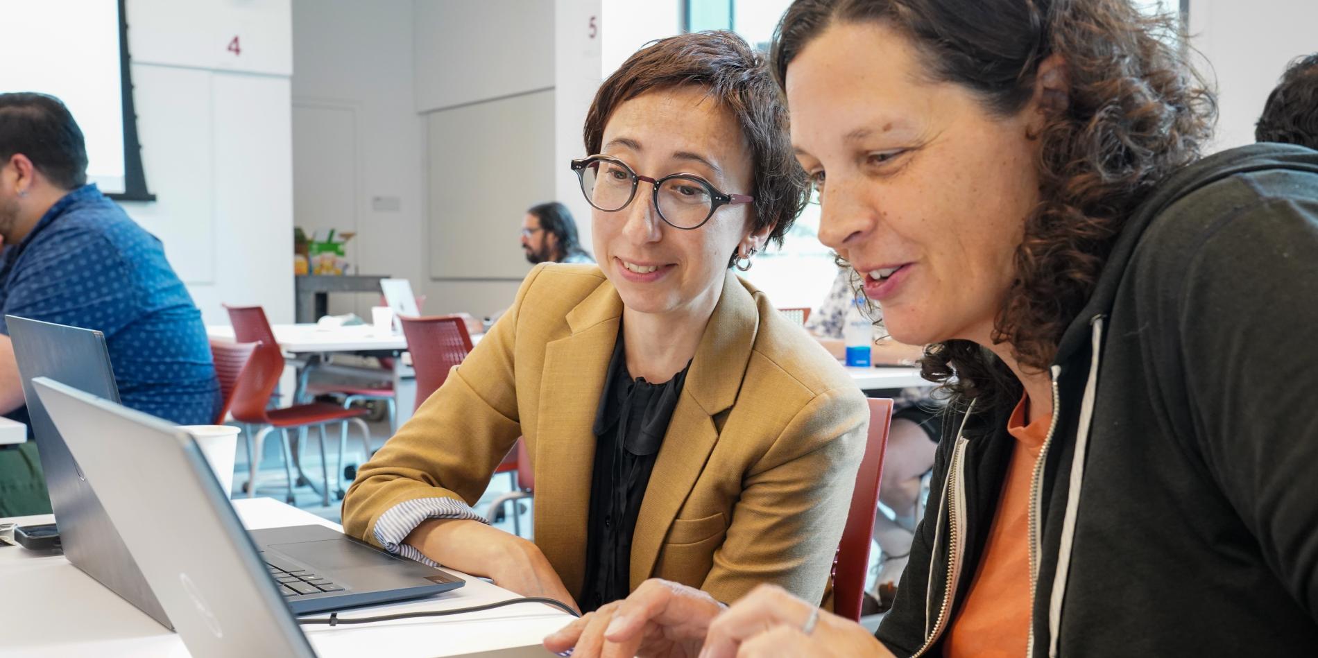 two women with brown hair type on two laptops and work 