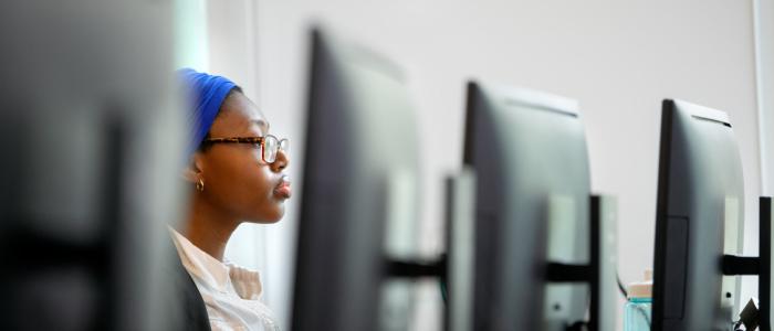 a black female student wearing a bright blue headpiece sits in between black computer monitors