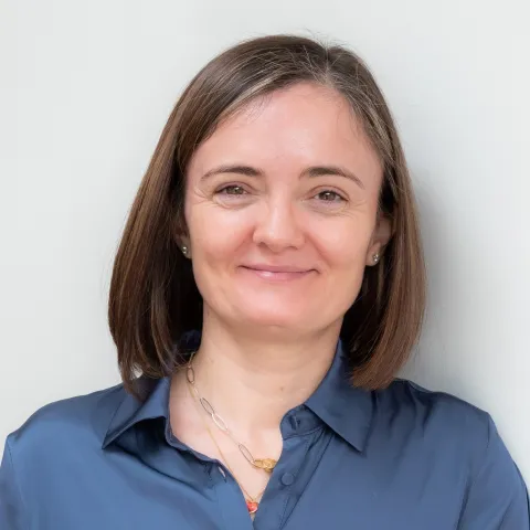 A photo of Alexandra Silva, a smiling woman with shoulder length brown hair and a blue shirt in front of a gray background
