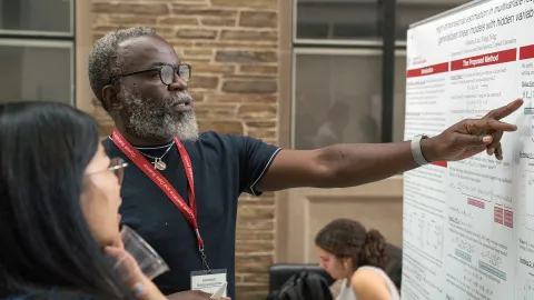A color photo of a man pointing at a poster while discussing it with a young woman.