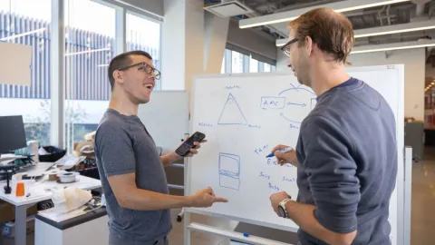 A color photo of 2 men discussing a project in front of a white board.