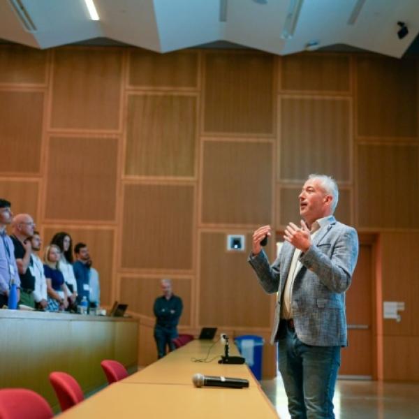A room with a man in a gray suit speaking to a group of people in the seats