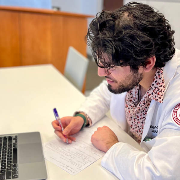 A medical student in a lab coat takes notes in front of a laptop