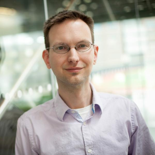 man with short, brown hair and glasses, leans against glass wall
