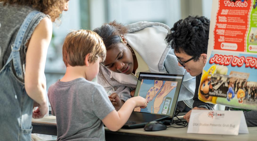 a young kid points to a game on a laptop as his mom and two college students look on.