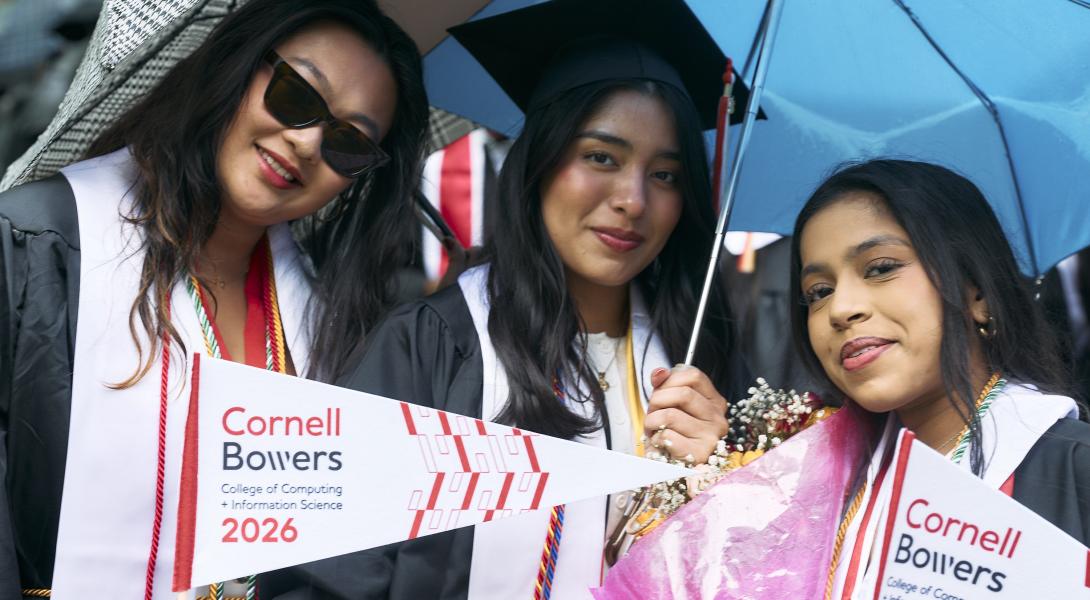 three young women wearing grad caps and waving white pennants smile under umbrellas.