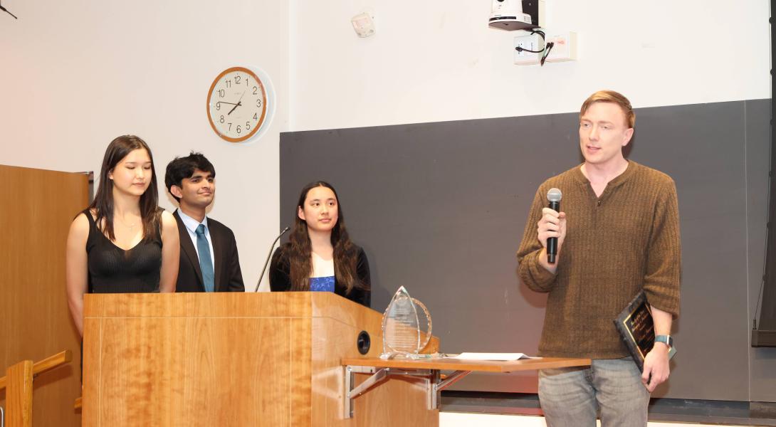 A color photo showing a man speaking into a microphone. With 3 people looking on.