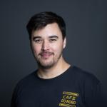 A photo of Michael Kim, a man with short black hair, a beard and mustache, in a black tshirt in front of a gray background
