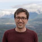 A photo of Nicholas Spooner, a smiling man with brown hair and glasses in a maroon sweater, in front of a mountain range.