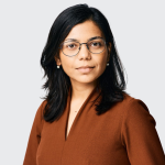 A photo of Rachee Singh, a woman with long dark hair and glasses in a brown sweater in front of a gray background