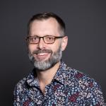 Portrait of man smiling at camera. he has on glasses and has a beard.