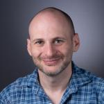A photo of Noah Stephens-Davidowitz, a smiling man with a shaved head, mustache and beard, and a blue plaid shirt in front of a gray background