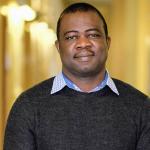 A photo of Owolabi Legunsen, a man with short black hair and mustache, in a black sweater in front of a gold background