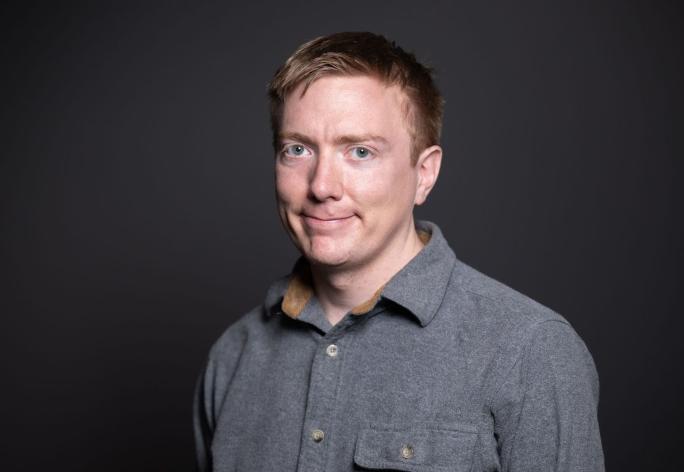 A photo of Adrian Sampson, a man with short dark-blond hair and a gray shirt in front of a gray background