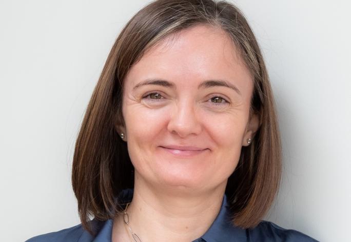 A photo of Alexandra Silva, a smiling woman with shoulder length brown hair and a blue shirt in front of a gray background
