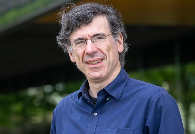 A portrait of Jon Kleinberg, a man with curly gray hair, glasses and a blue shirt, in front of Gates Hall