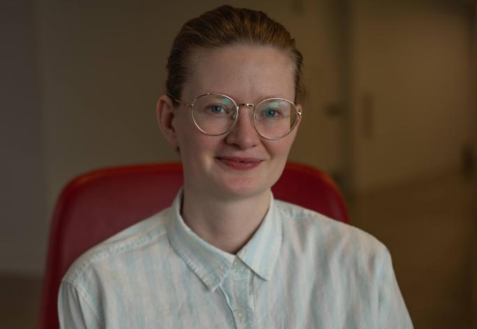 A photo of Sarah Dean, a woman with glasses and blond hair pulled back and a white shirt, sitting in a red chair.