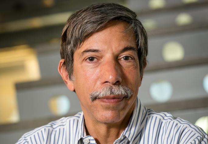 A photo of Fred Schneider, a man with dark gray hair and a mustache, in a striped shirt in front of a staircase
