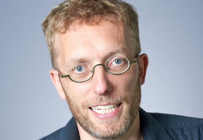 A photo of Kilian Weinberger, a man with curly blond hair, glasses, a beard and mustache, in a gray shirt in front of a gray background