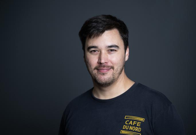 A photo of Michael Kim, a man with short black hair, a beard and mustache, in a black tshirt in front of a gray background