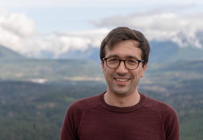 A photo of Nicholas Spooner, a smiling man with brown hair and glasses in a maroon sweater, in front of a mountain range.