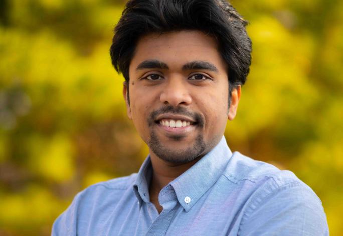 A photo of Saikat Dutta, a man with dark brown hair and a beard, wearing a blue shirt in front of a leafy background