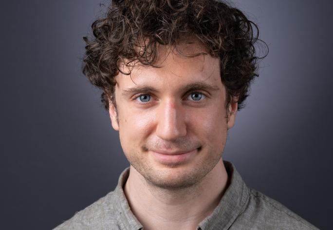 A photo of Kevin Ellis, a man with curly brown hair wearing a beige-gray shirt in front of a gray backround.