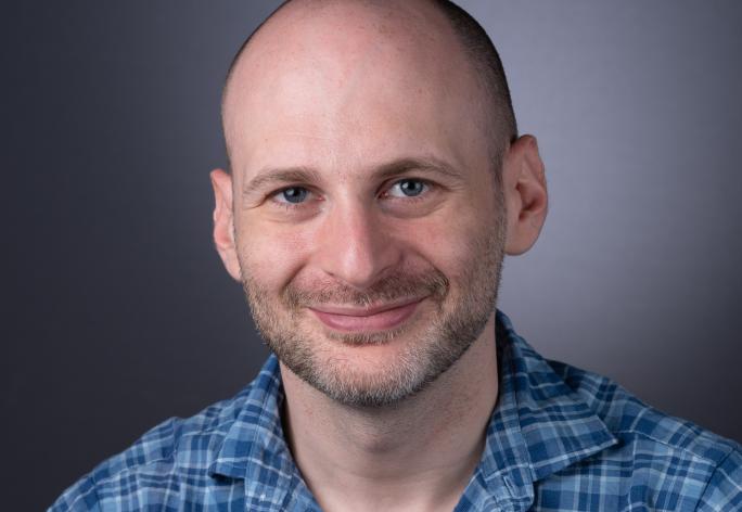 A photo of Noah Stephens-Davidowitz, a smiling man with a shaved head, mustache and beard, and a blue plaid shirt in front of a gray background