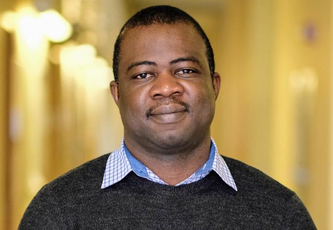 A photo of Owolabi Legunsen, a man with short black hair and mustache, in a black sweater in front of a gold background