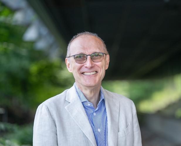 Man with glasses, short hair, blue shirt and tan jacket smiling at the camera