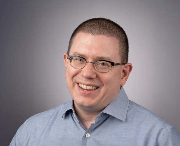 A color photo of Michael Clarkson, a smiling man with a buzzed head, glasses and a gray shirt in front of a gray background