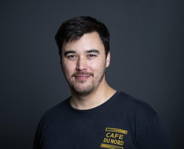 A photo of Michael Kim, a man with short black hair, a beard and mustache, in a black tshirt in front of a gray background