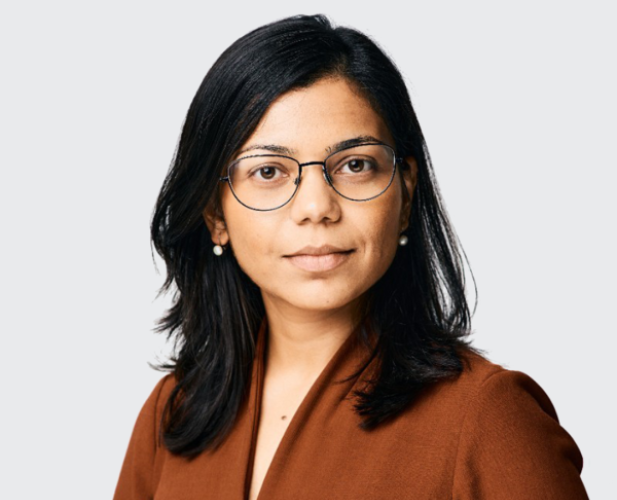 A photo of Rachee Singh, a woman with long dark hair and glasses in a brown sweater in front of a gray background
