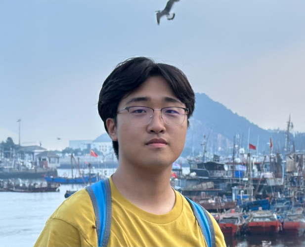 A color photo of a man with glasses in front a bay with boats.