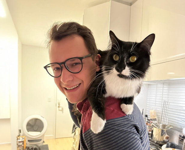 A color photo of a man with glasses smiling for a photo while a black and white cat sits on his shoulders.