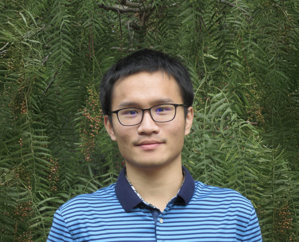 A color photo of a man smiling for a photo in front of green trees.