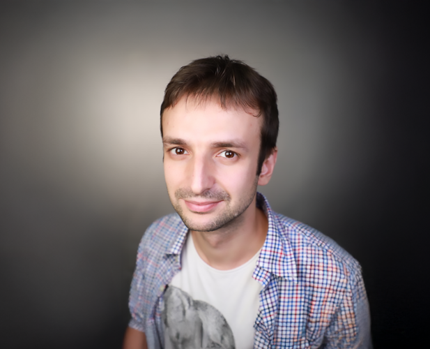 A headshot of a man with short brown hair, wearing a tshirt and button-down