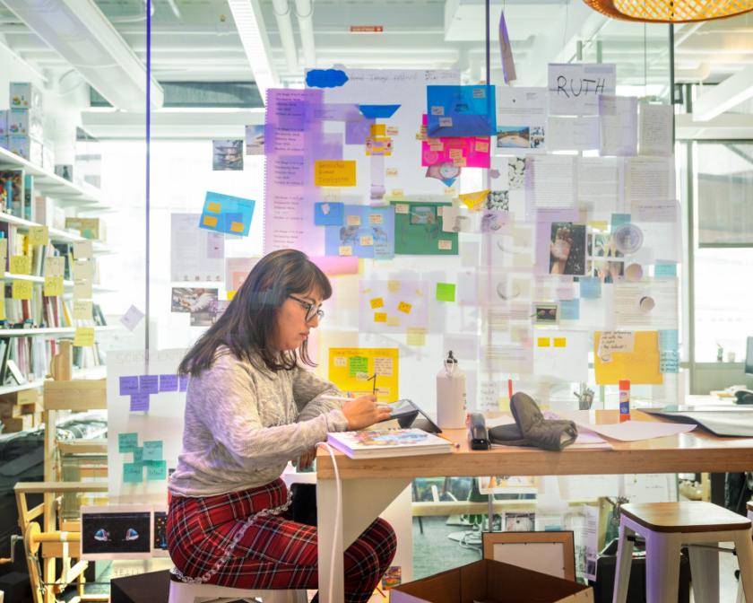A color photo of a woman working at a desk, surrounded by notes.