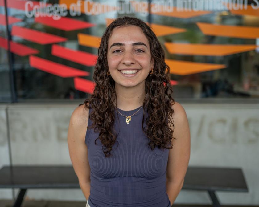 a female student with tan skin and long brown curly hair wearing a dark purple gray tank top poses in front of windows painted red and orange