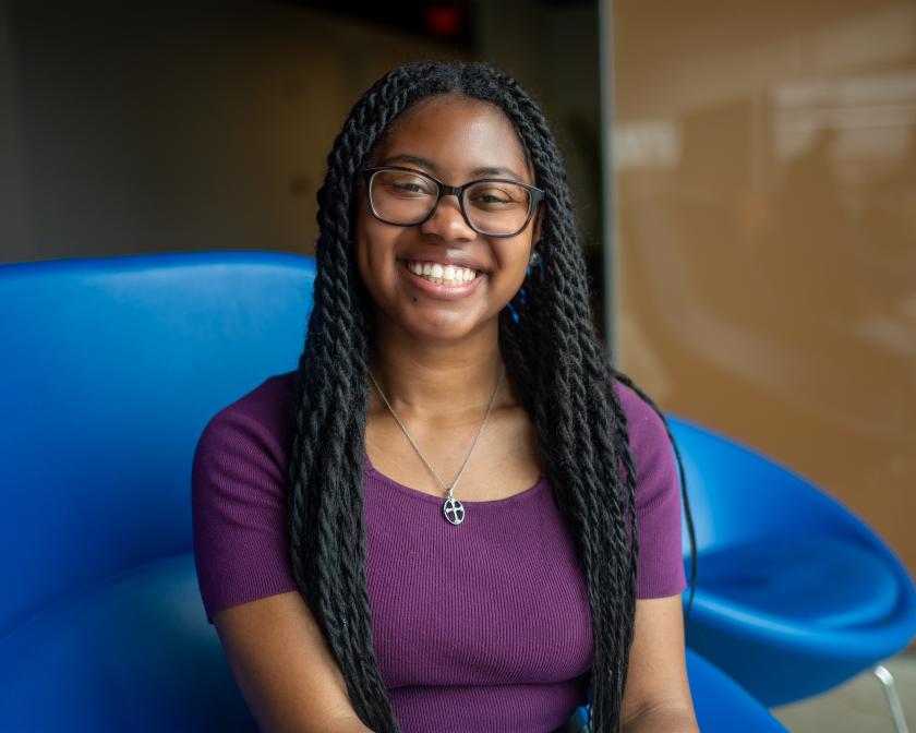 a young black female student with long black hair in braids wears a purple shirt and sits on a bright blue chair