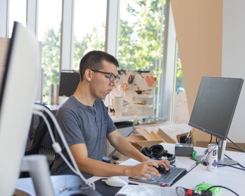 a male white student with glasses sits and types on a keyboard in front of a black monitor