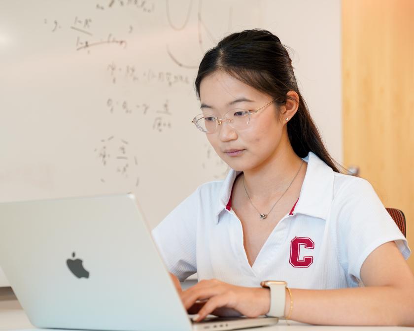 A color photo of a woman with glasses working on a laptop.