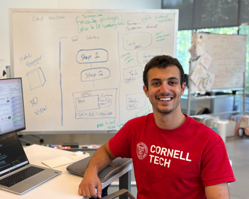 A color photo of a man smiling for a photo at his workstation.
