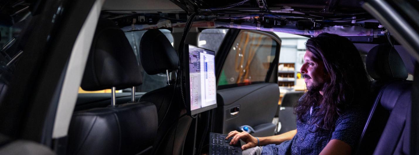 inside of a dark car a man with long dark hair sits in the backset looking at a computer screen
