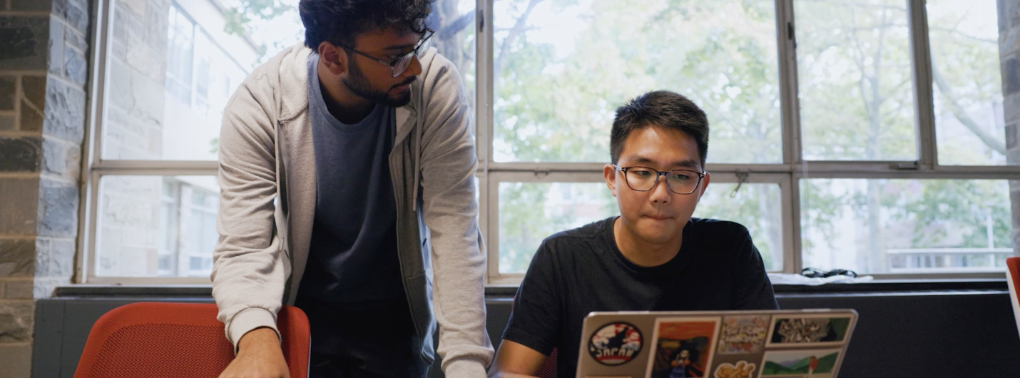 two students (one standing, one sitting) look at computer laptop