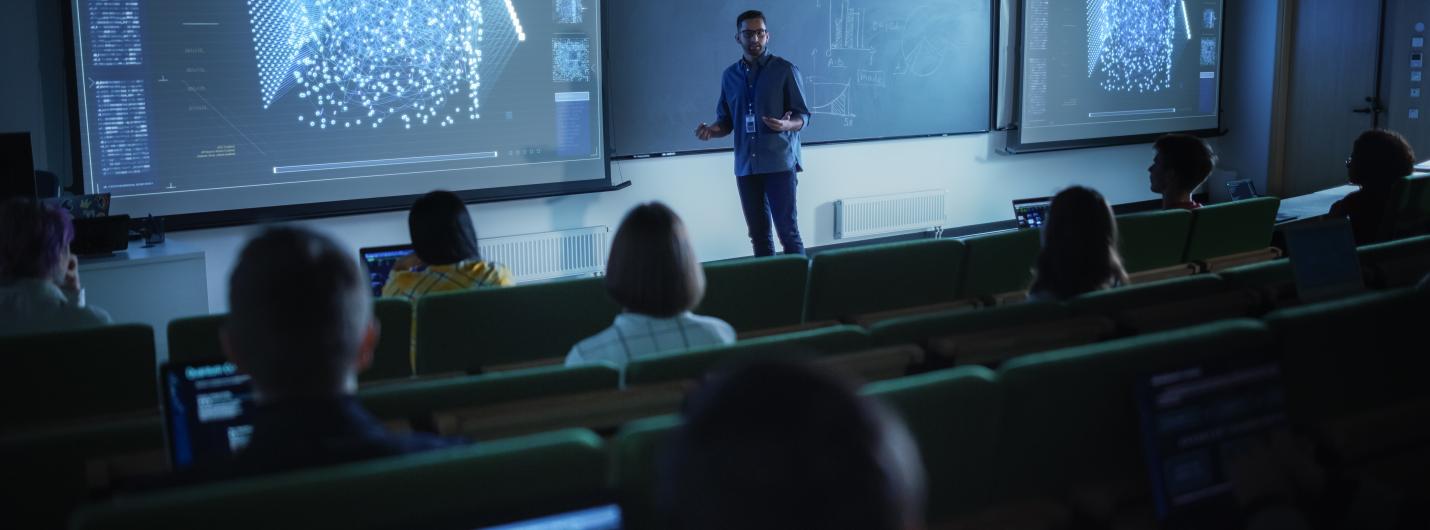 a person presents in front of two large screens with blue charts and data, people sit in audience 
